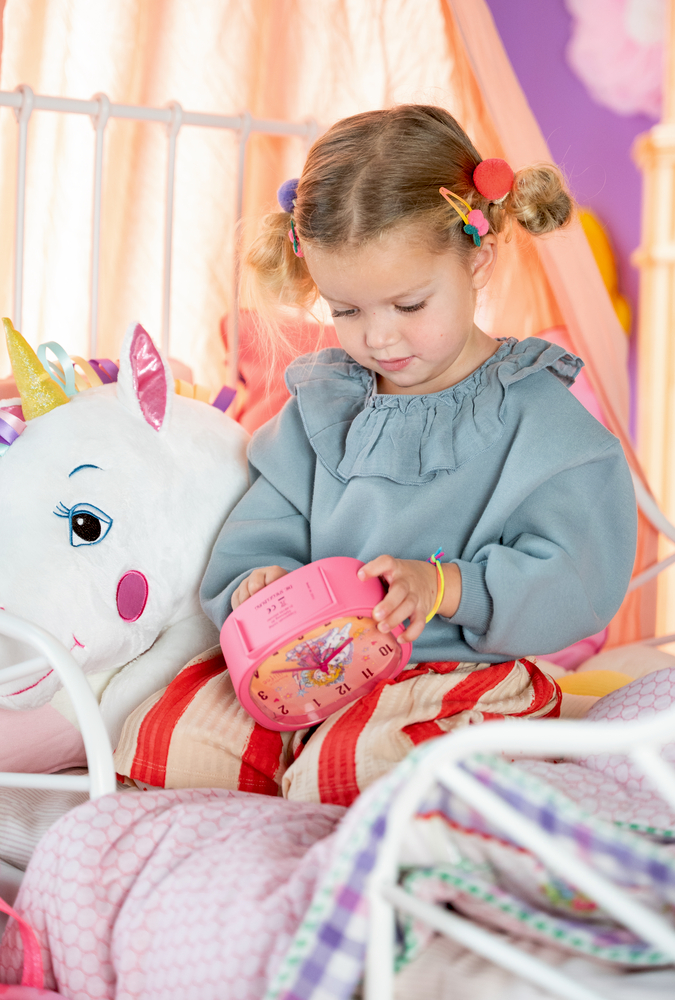 Mädchen sitzt auf Bett mit Einhornplüschtier, schaut auf pinkfarbenen Wecker, buntes Zimmer.