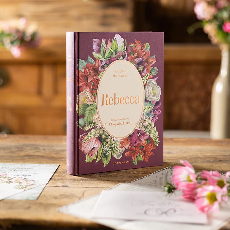 Das weinrote Buch "Rebecca" auf einem Tisch, dekoriert mit Blumen.