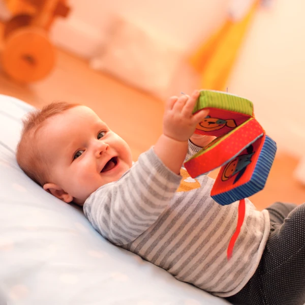 Lachendes Baby liegt auf dem Rücken und hält ein Stoffbuch in den Händen.