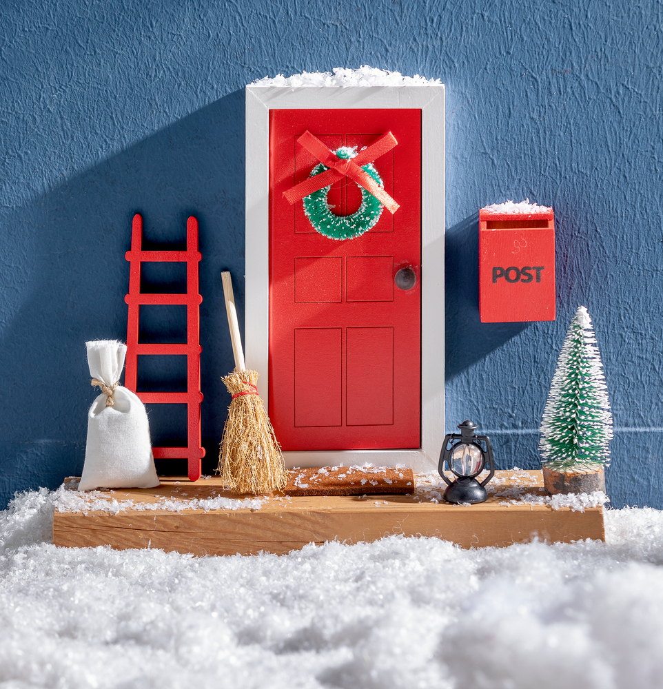 Kleine rote Tür mit Kranz, Leiter, Briefkasten und schneebedecktem Dekor auf einer verschneiten Oberfläche.