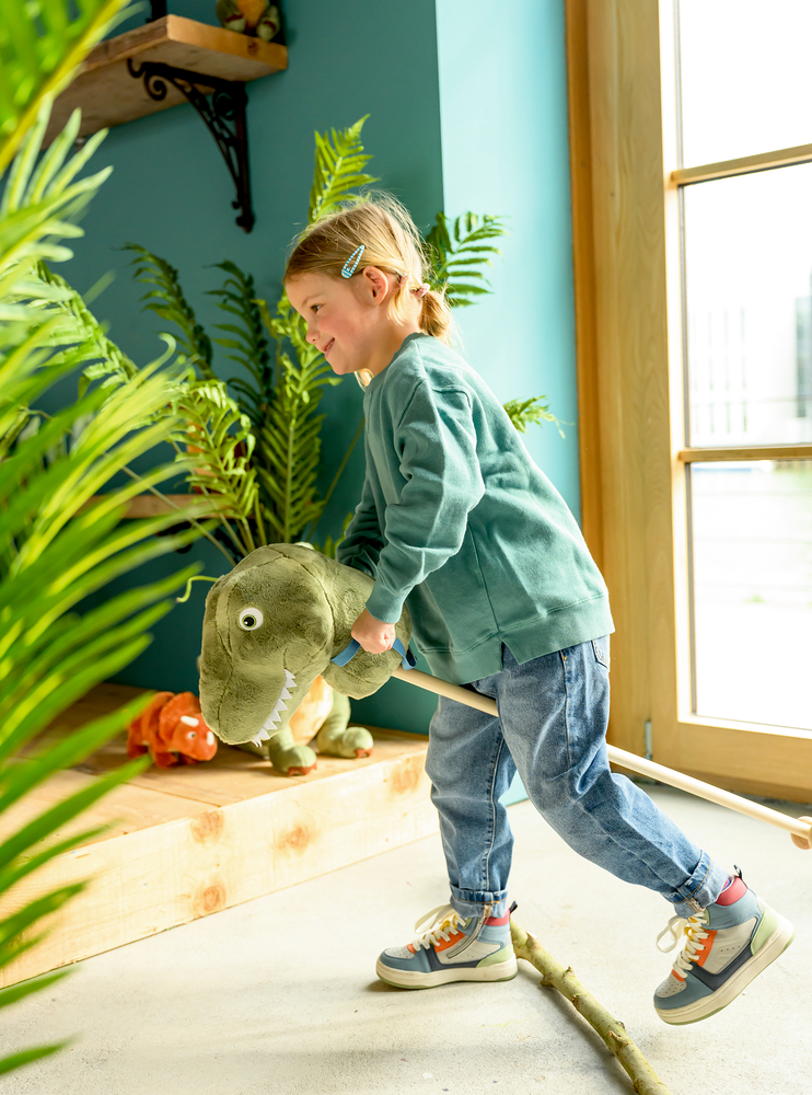 Lächelndes Kind reitet drinnen auf einem Spielzeug-Dinosaurierstab, umgeben von grünen Pflanzen und Holzdekor.