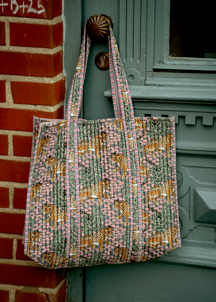 Eine Stofftasche mit Leoparden- und Blumenmuster hängt an einer blaugrünen Tür neben einer Backsteinwand.