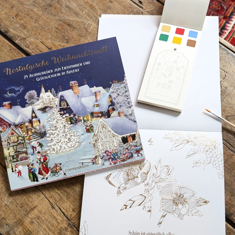 Weihnachtliche Malblöcke im geschlossenen und im offenen Zustand sowie Pinsel und Farbkarte auf einem alten Tisch.