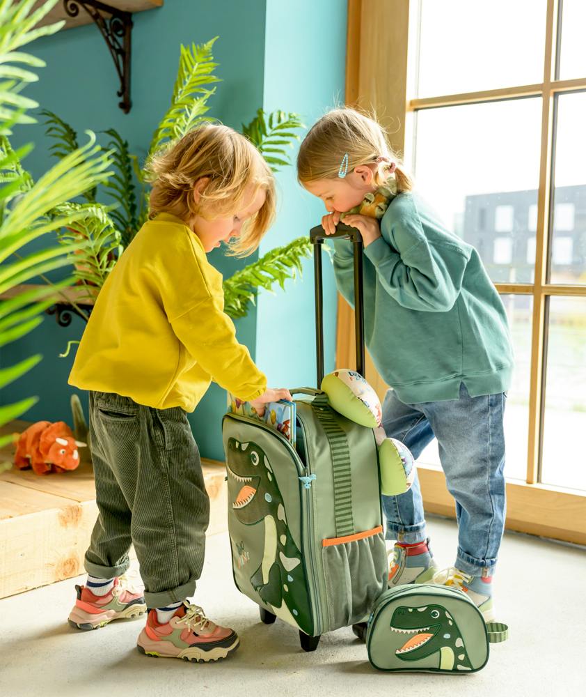 Zwei Kinder packen einen grünen Dinosauriertrolley an einem Fenster, umgeben von Pflanzen und Spielzeug.