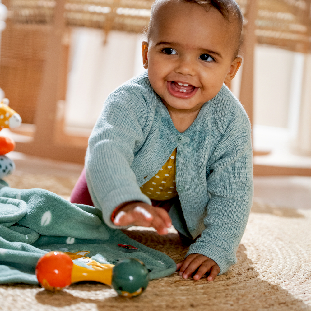 Baby im blauen Pullover krabbelt auf Teppich und greift nach buntem Spielzeug auf grünem Tuch.