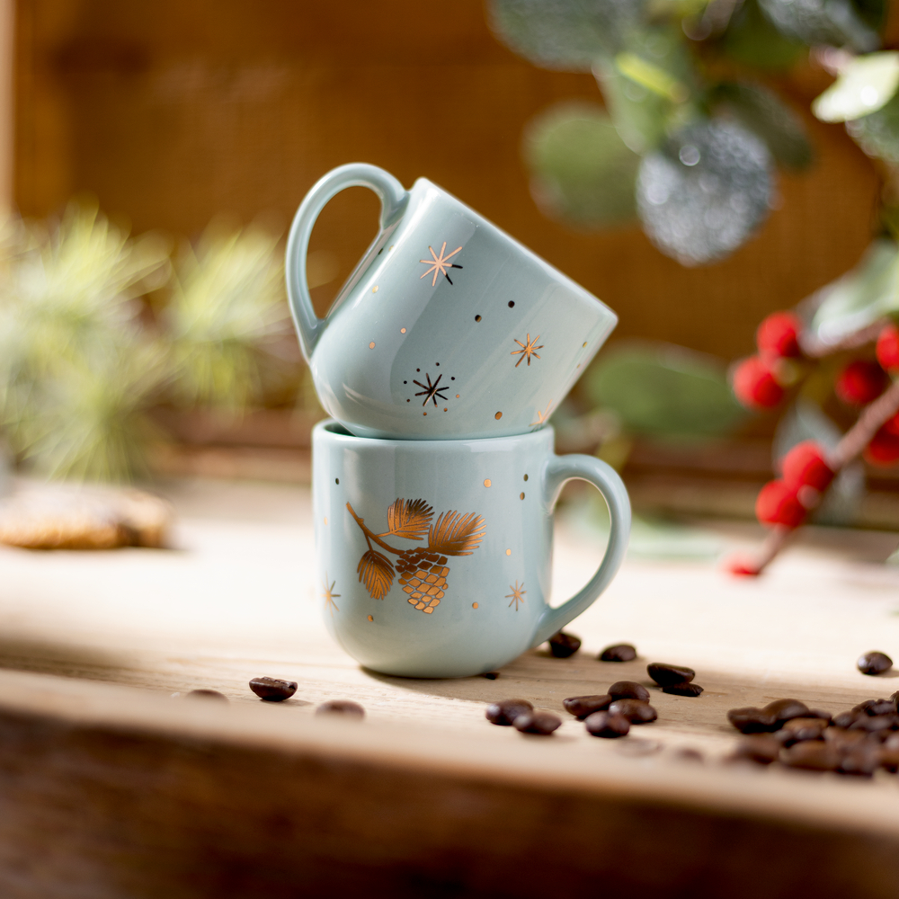 Zwei salbeifarbene Tassen mit Goldmotiven auf einem weihnachtlichen Holztisch. 