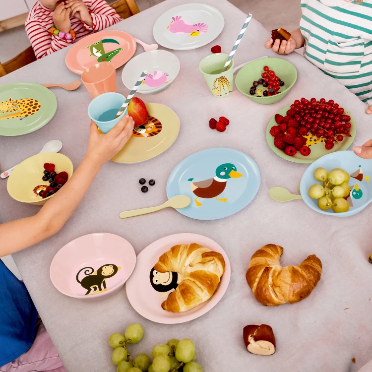 Ausschnitt eines Tischs mit buntem Kindergeschirr, Obst, Croissants und Kinderhänden.