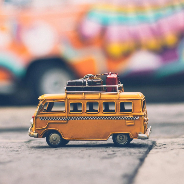Ein kleiner gelber Spielzeug-Bus mi bunten Koffern auf dem Dach vor einem bunten verschwommenen Hintergrund.