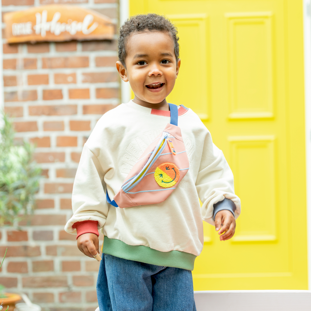 Kind mit weißem Pullover und rosa Tasche steht vor einer gelben Tür. Das Kind lächelt.