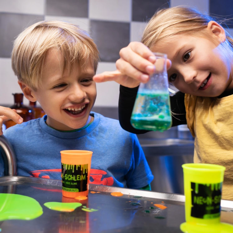 Kinder betrachten ein Reagenzglas mit blauer Flüssigkeit. Auf einem Tisch stehen neonfarbene Dosen mit herauslaufender, greller Substanz.