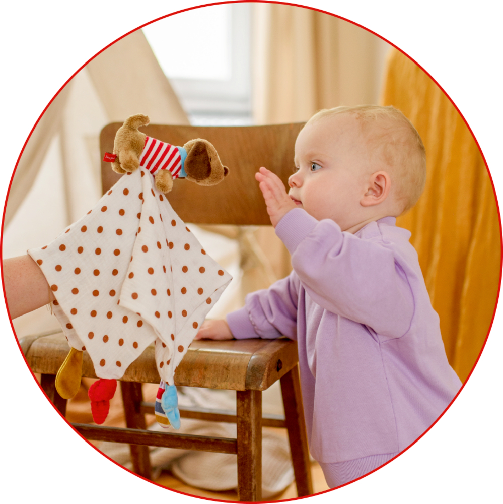 Baby im lila Pullover greift nach einer braunen Hundehandpuppe mit gepunktetem Tuch.