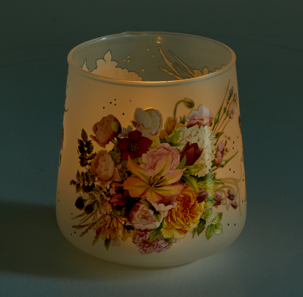 Auf einer dunklen Oberfläche steht ein beleuchtetes Kerzenglas, geschmückt mit bunten Blumen.