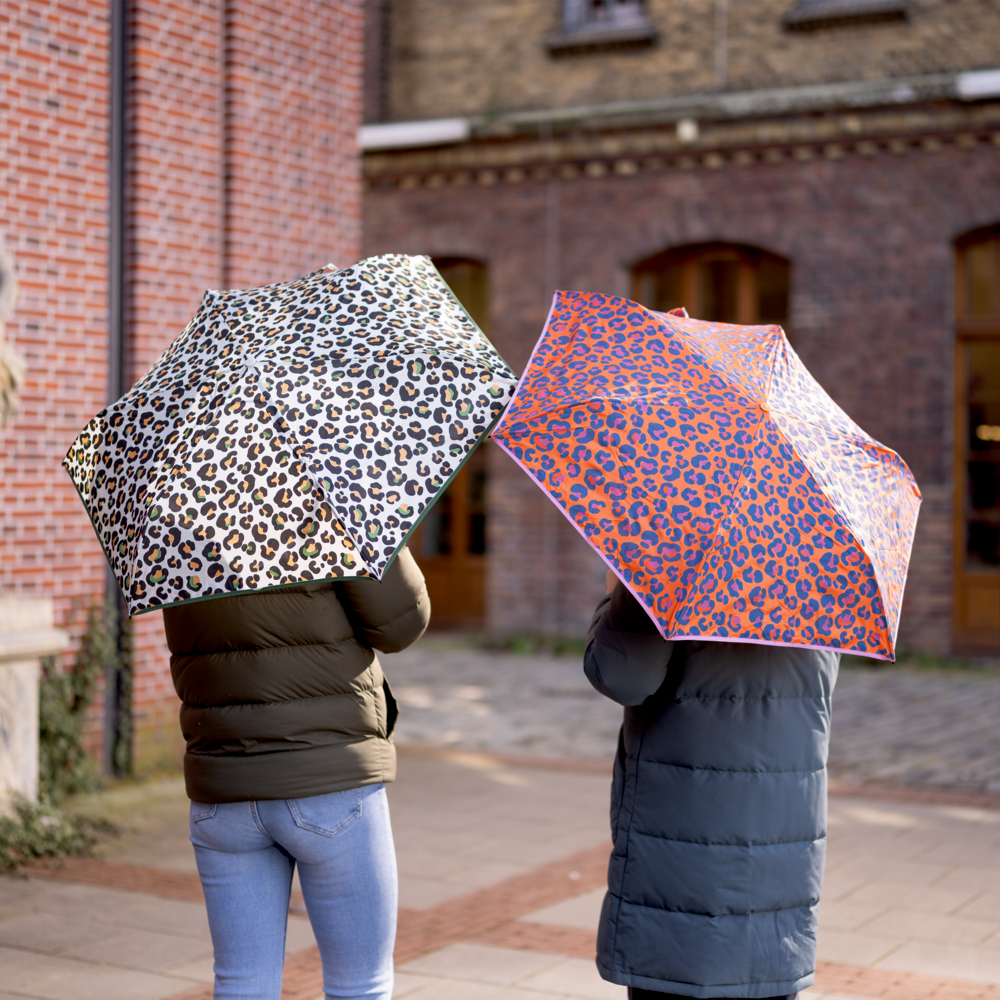 Zwei Menschen mit Regenschirmen im Leopardenmuster stehen unter einem bewölkten Himmel vor Backsteingebäuden.