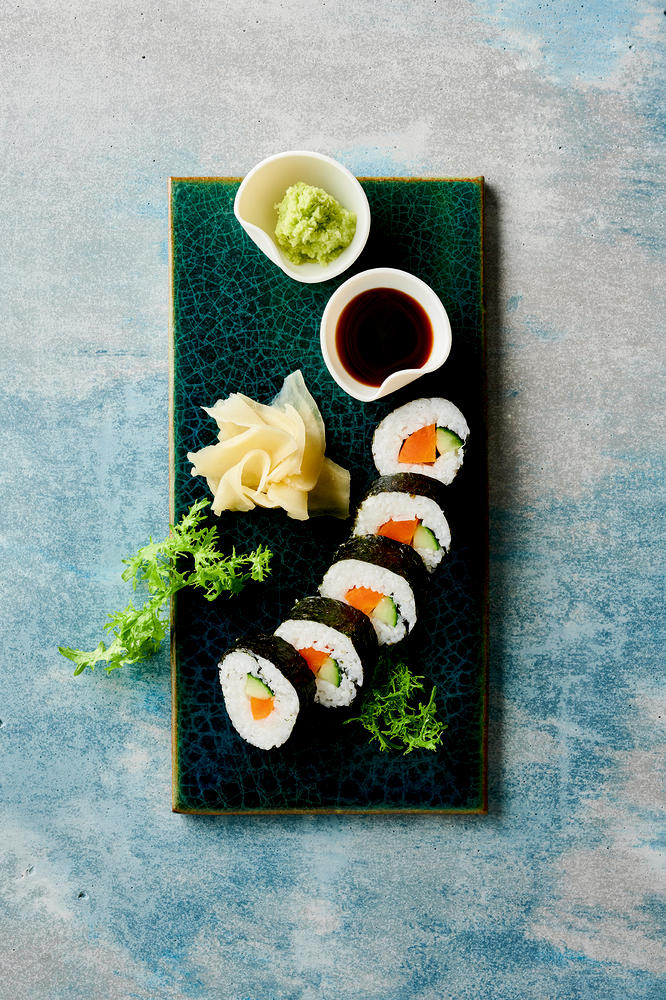 Sechs Gemüse-Sushi-Rollen mit Wasabi, Sojasauce, eingelegtem Ingwer und Gemüse, serviert auf einem rechteckigen Teller.