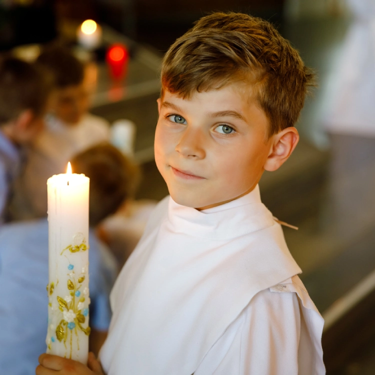 Junge mit weißer Kommunionskleidung hält eine angezündete Kerze in den Händen.