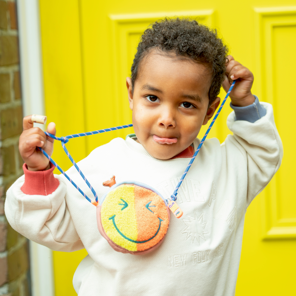 Ein Kind im weißen Pullover hält einen Smiley-Brustbeutel, streckt die Zunge heraus und steht vor einer gelben Tür.