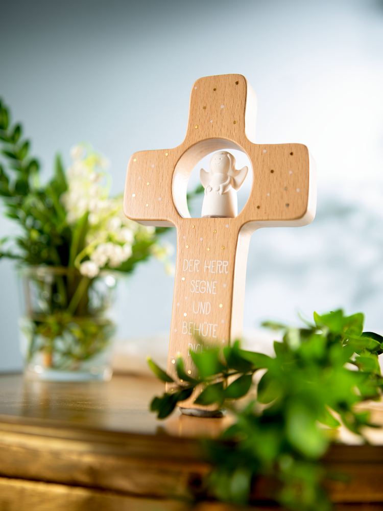 Holzkreuz mit kleinem weißen Engel als Mittelstück, umgeben von grünen Pflanzen stehend auf einer Holzoberfläche.