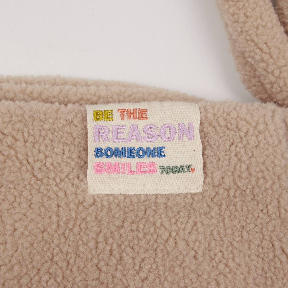 Beiges Fleece mit einem bunten Etikett mit der Aufschrift „Seien Sie heute der Grund, warum jemand lächelt.“.