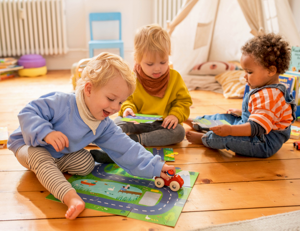 Drei Kleinkinder spielen mit Spielzeugautos und Matten auf einem Holzboden in einem gemütlichen, hellen Raum.