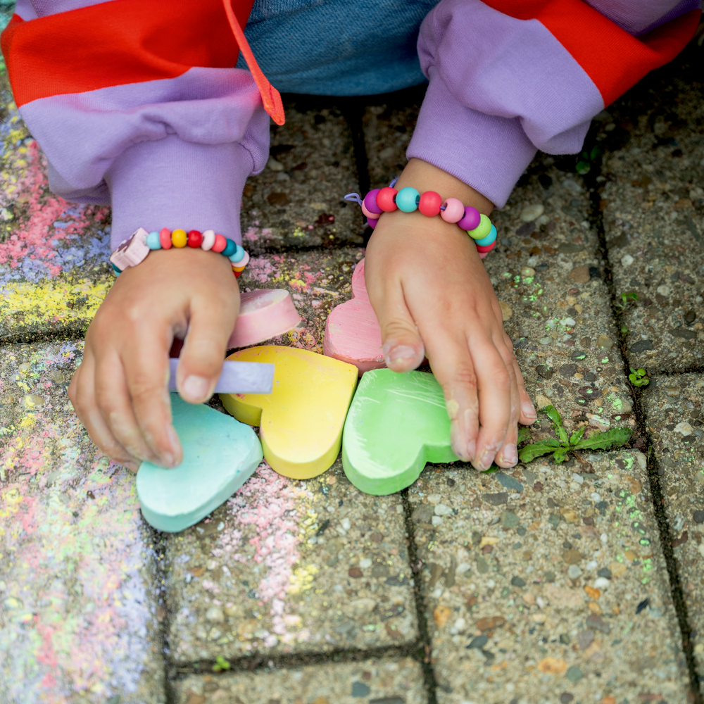 Kind malt auf Gehweg mit Herz-Kreide und trägt bunte Perlenarmbänder.