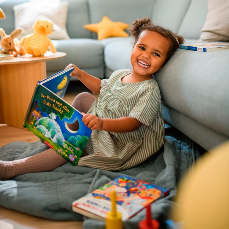 Mädchen mit Bilderbuch auf einem Teppich lacht in die Kamera.