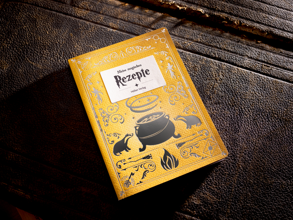 Ein gelbes Rezeptbuch mit Tier- und Kesselillustrationen auf einem rustikalen Holztisch.