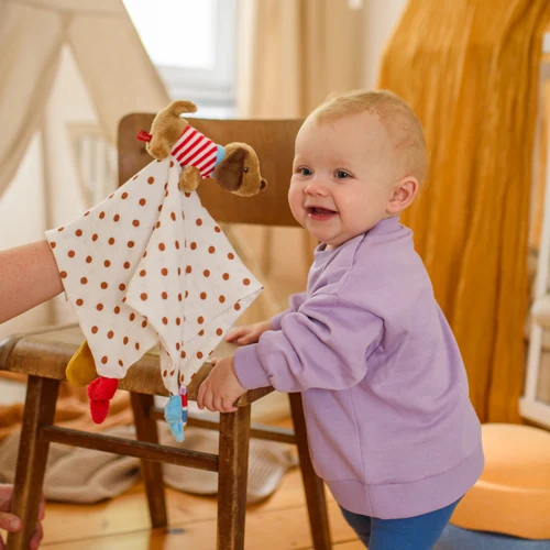 Eine Hand hält einem lachenden Baby ein Schnuffeltuch mit einem Hündchen hin.