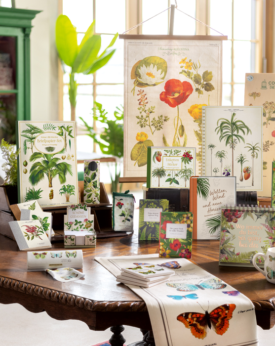 Zusammenstellung verschiedener Kalender, Büchlein, Handtücher und Karten mit Naturmotiven auf einem dunkelbraunem Tisch vor einem großen Fenster