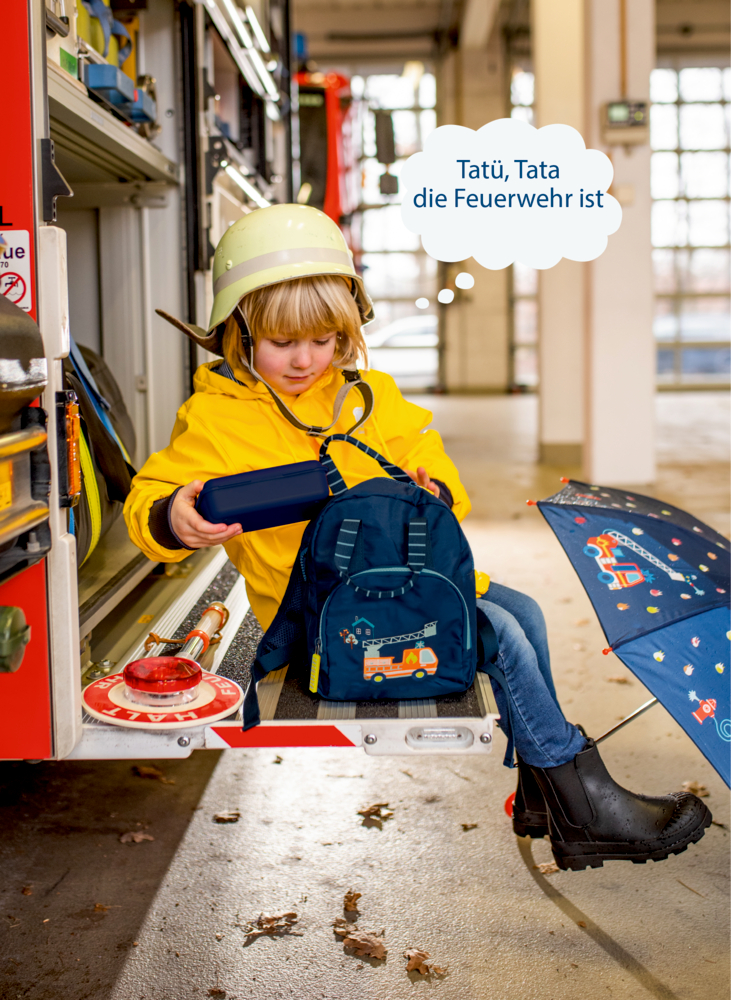 Ein Kind im gelben Regenmantel sitzt mit Feuerwehrhelm und Lunchbox in einem Feuerwehrauto.