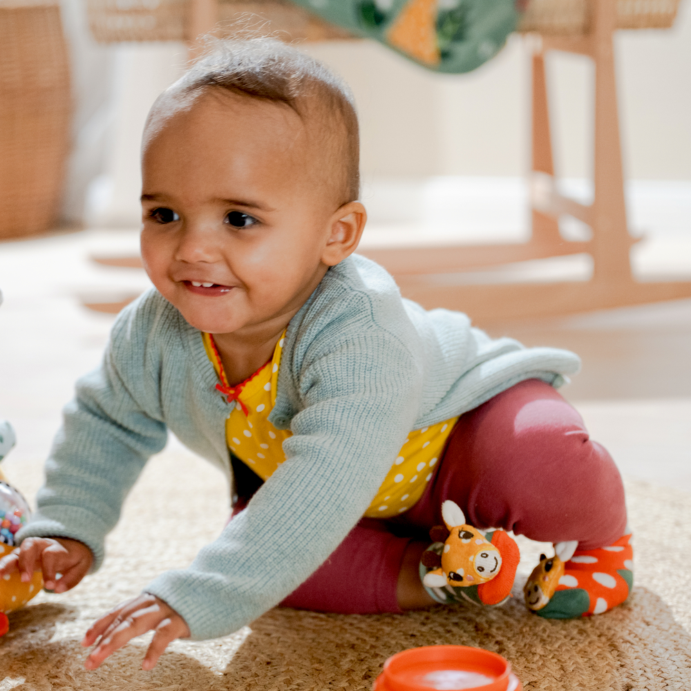 Baby lächelt, krabbelt auf Teppich und hat bunte Giraffensöckchen an.