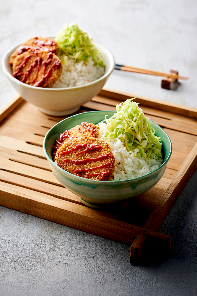 Zwei Schüsseln Reis mit panierten Koteletts, geschnetzeltem Kohl und roter Soße auf einem Holztablett.