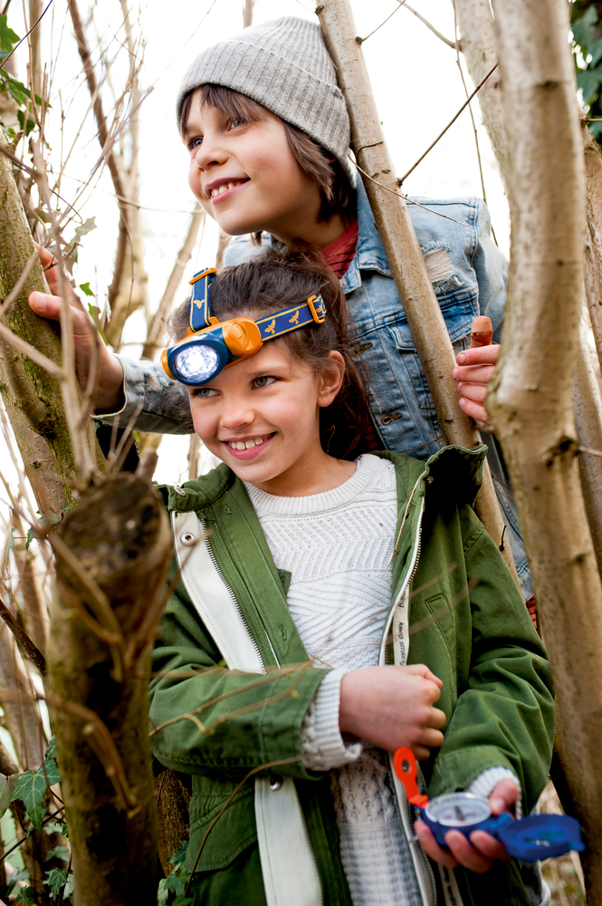 Zwei lächelnde Kinder erkunden den Wald – eines mit einer Stirnlampe, das andere mit einem Kompass.