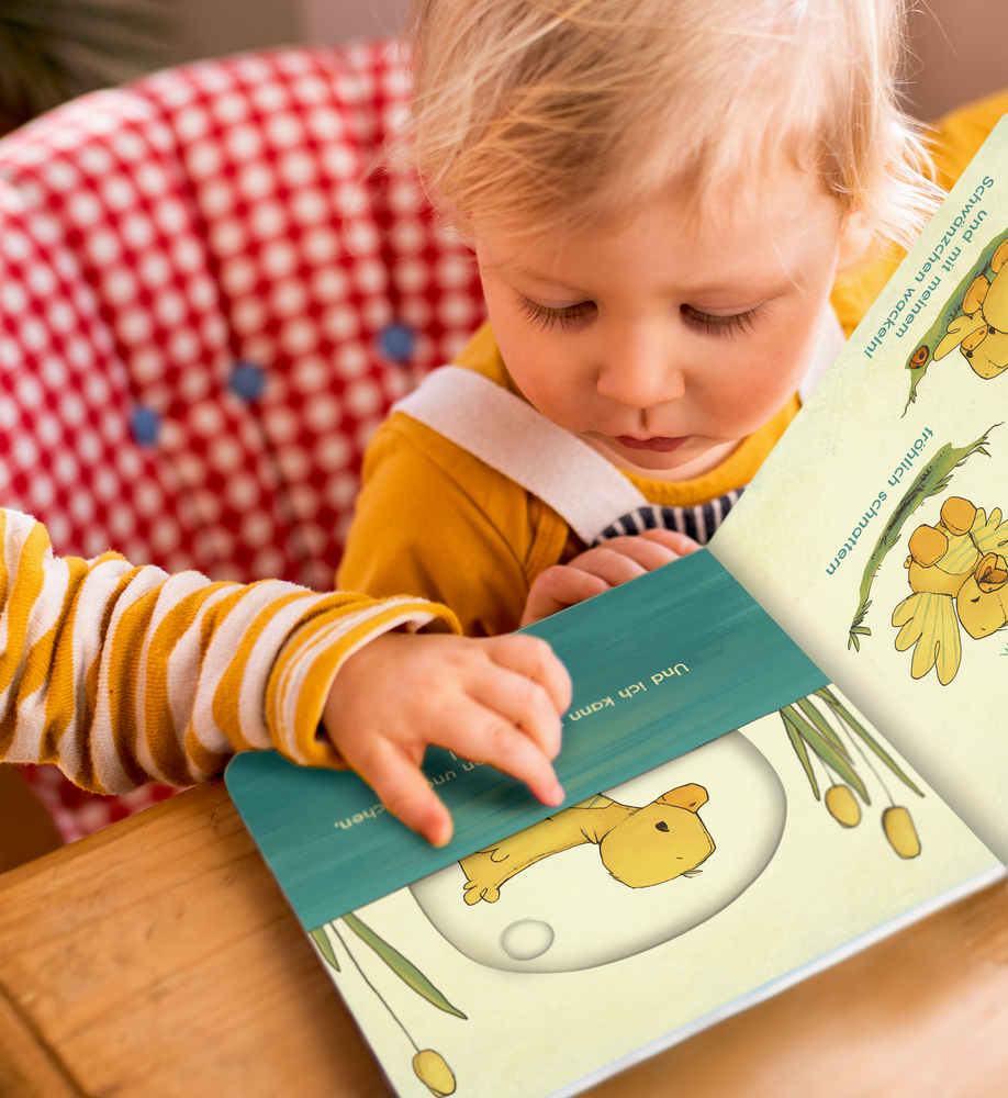 Ein Kleinkind in Gelb sitzt an einem Tisch und liest ein Bilderbuch mit Tierillustrationen.