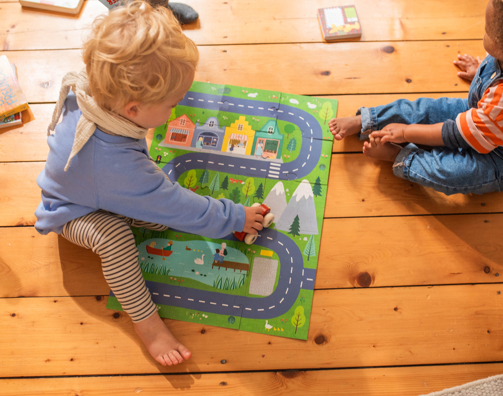 Zwei kleine Kinder spielen auf einem Holzboden mit einer bunten Spielmatte mit Straßen und einer Stadt.