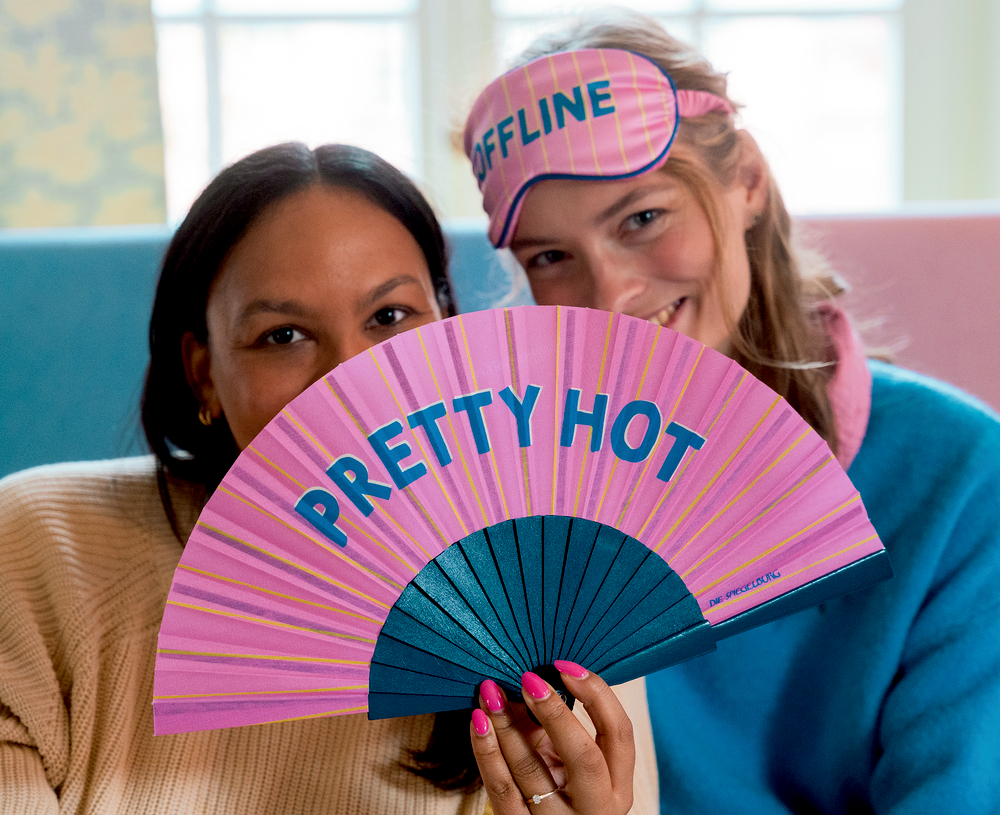 Zwei Frauen lächeln, eine hält einen pinken Fächer mit der Aufschrift PRETTY HOT, die andere trägt eine OFFLINE Schlafmaske.