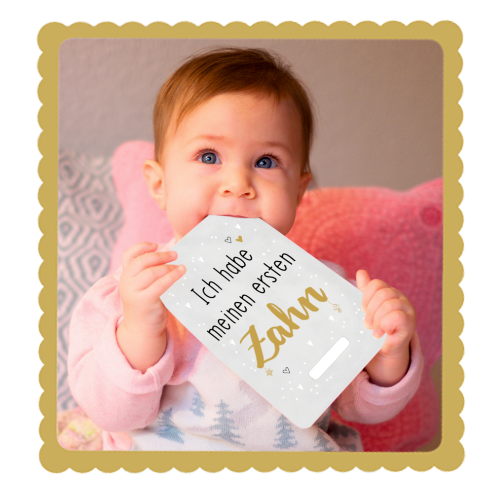 Baby lächelt sitzend auf einem rosa Kissen und hält eine Karte mit der Aufschrift „Ich habe meinen ersten Zahn“.