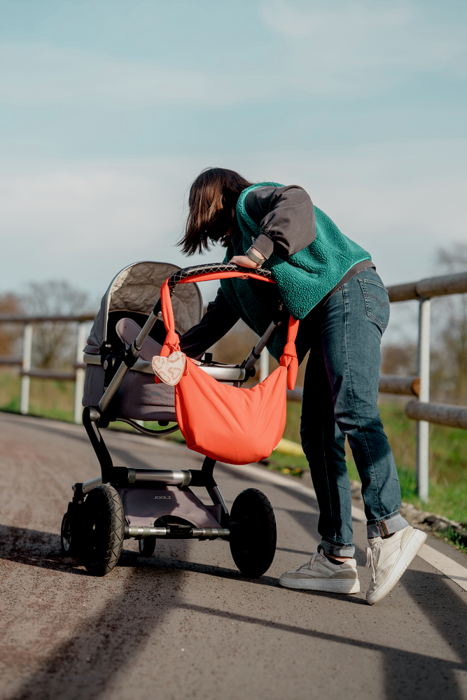 Person beugt sich über einen grauen Kinderwagen mit orangefarbener Tasche am Griff auf einem Weg ins Freie.