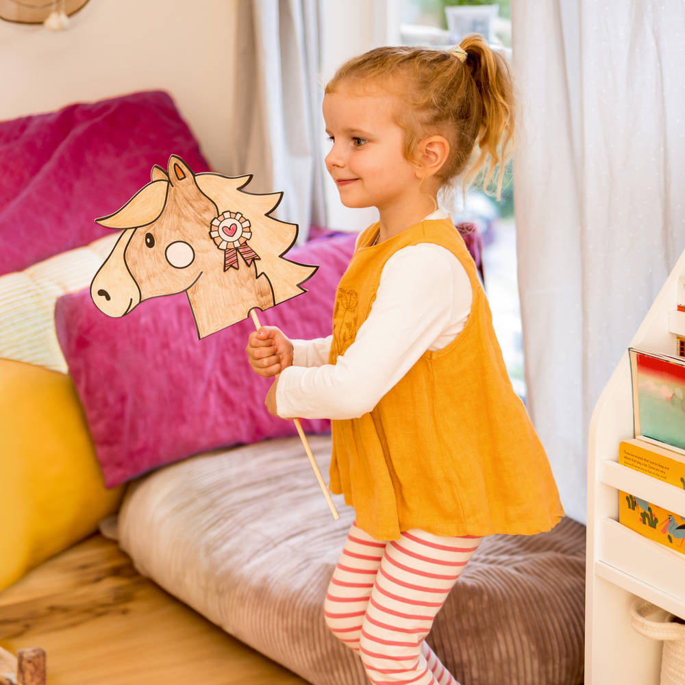 Ein lächelndes Mädchen in einem gelben Kleid spielt drinnen und hält einen Stock mit einem Pferdekopf aus Papier.