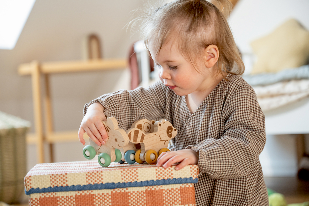 Kind spielt mit drei hölzernen Tierfiguren auf Rädern auf einer bunten Box.