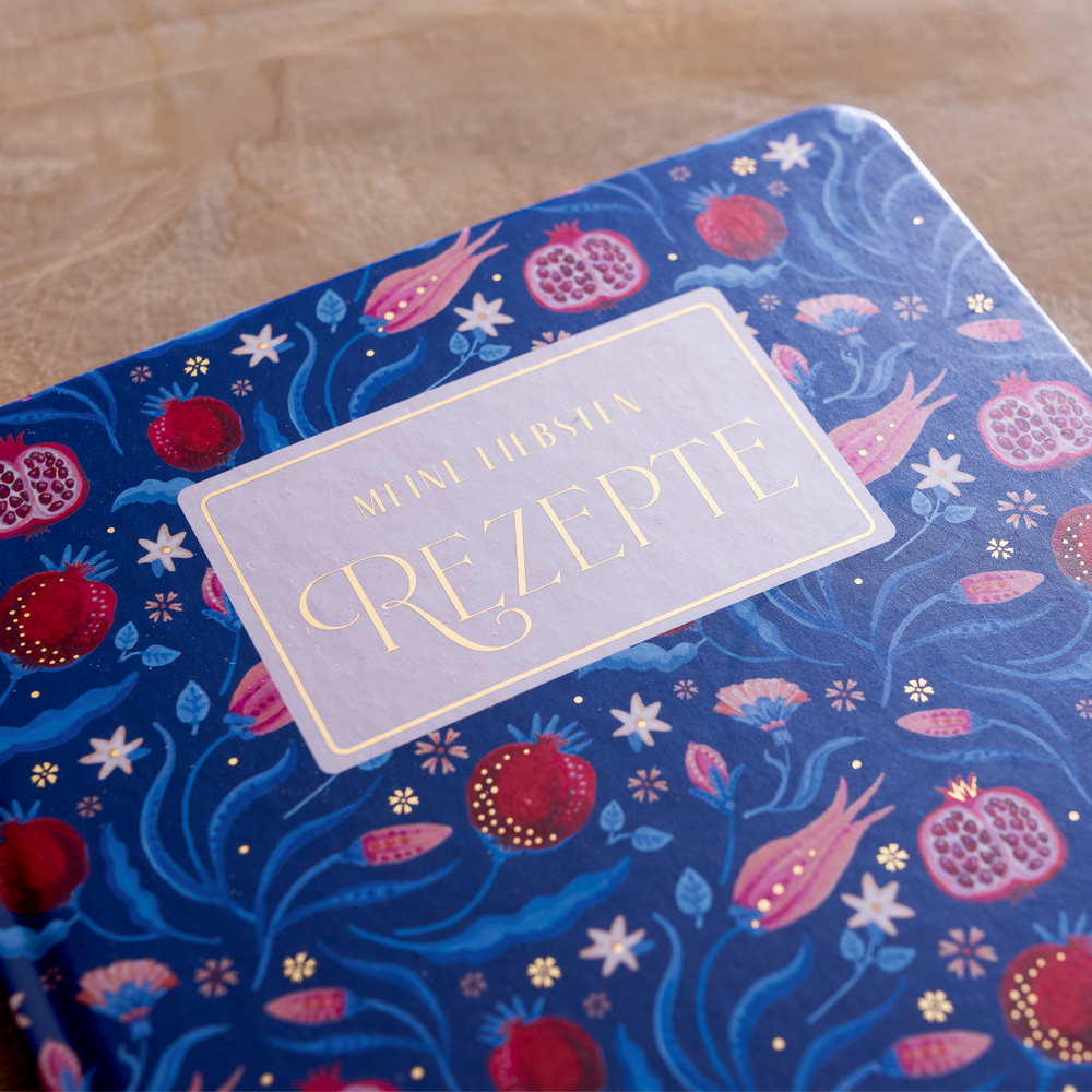 Blaues Rezeptbuch mit floralen und granatapfelartigen Mustern, goldener Schrift „Meine liebsten Rezepte“ auf dem Umschlag.