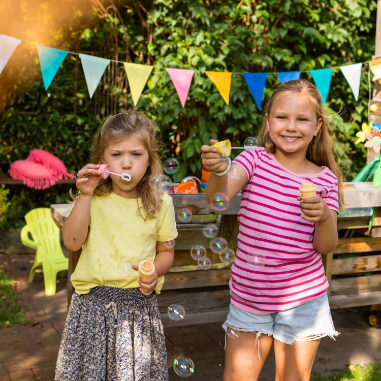 Zwei Mädchen im Garten mit Wimpelkette machen Seifenblasen.