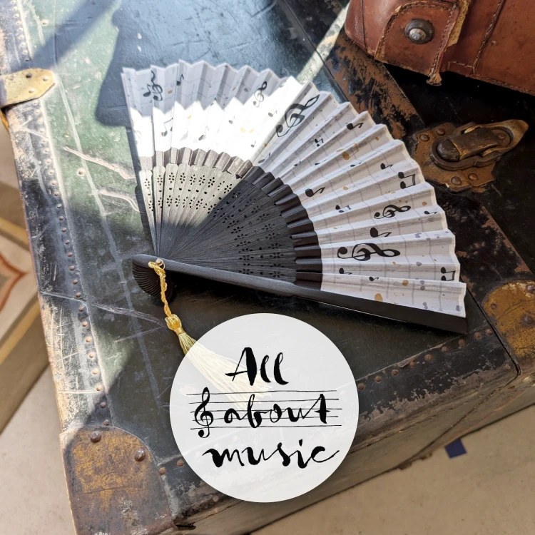 Schwarz-weißer Fächer mit Musikmotiven liegt auf einem alten Koffer im Sonnenschein.