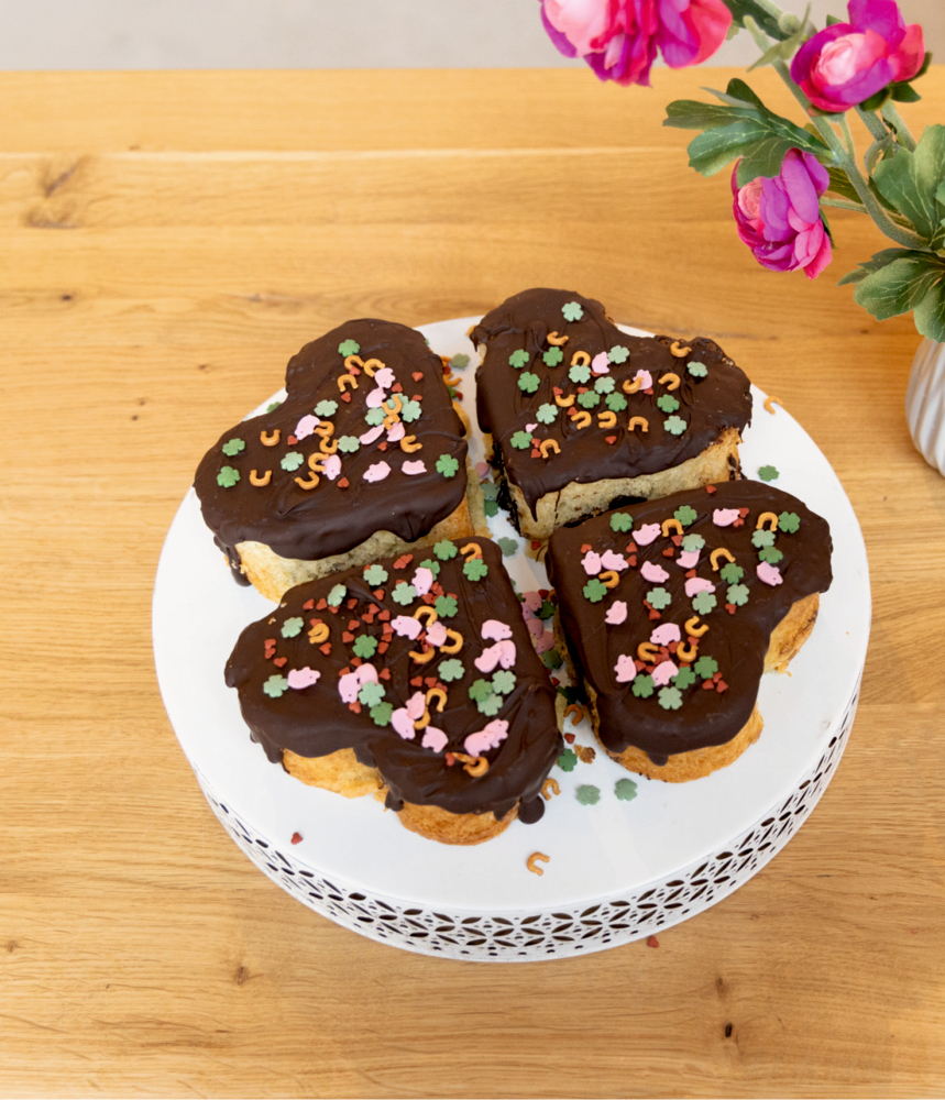 Schokoladenkuchen in Kleeblattform mit Streuseln auf einem weißen Teller, neben rosa Blumen auf einem Holztisch.
