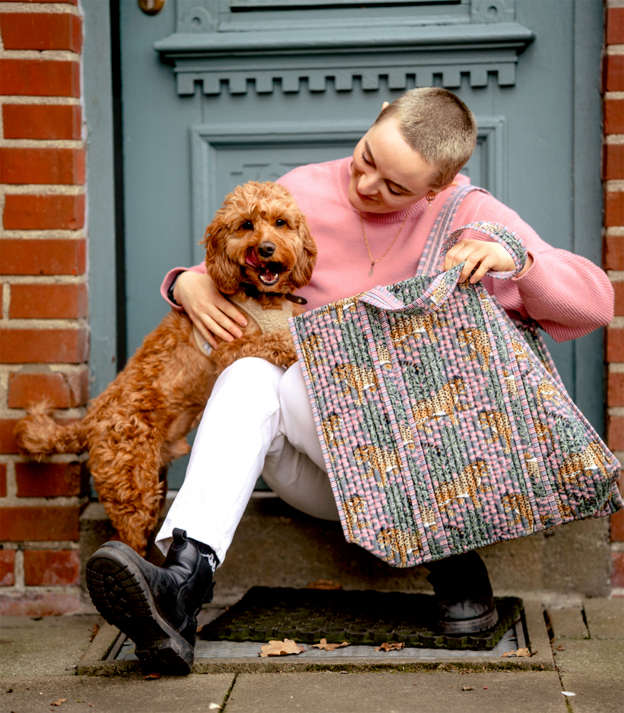 Person mit rosa Pullover sitzt lächelnd auf der Türschwelle, hält eine gemusterte Stofftasche in der Hand und umarmt einen fröhlichen braunen Hund.