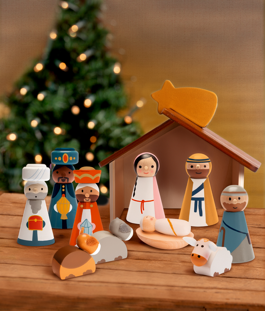 Holz-Krippenset mit Figuren, Tieren und Stall, im Hintergrund ein unscharfer Weihnachtsbaum mit Lichtern auf Produktverpackung.
