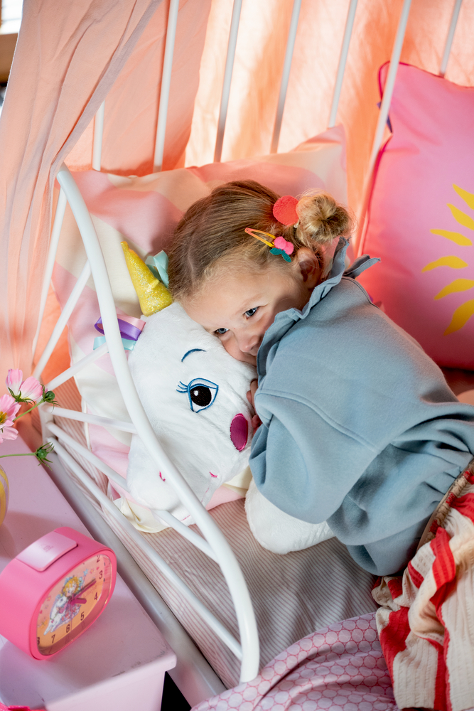 Ein Mädchen hält ein Einhorn-Kissen im Bett, lächelt, trägt bunte Haarspangen.