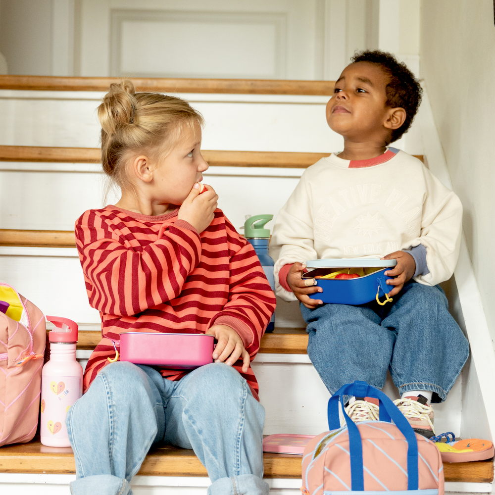 Zwei Kinder sitzen auf Treppenstufen. Sie essen aus Brotdosen. Taschen und Trinkflaschen stehen daneben.