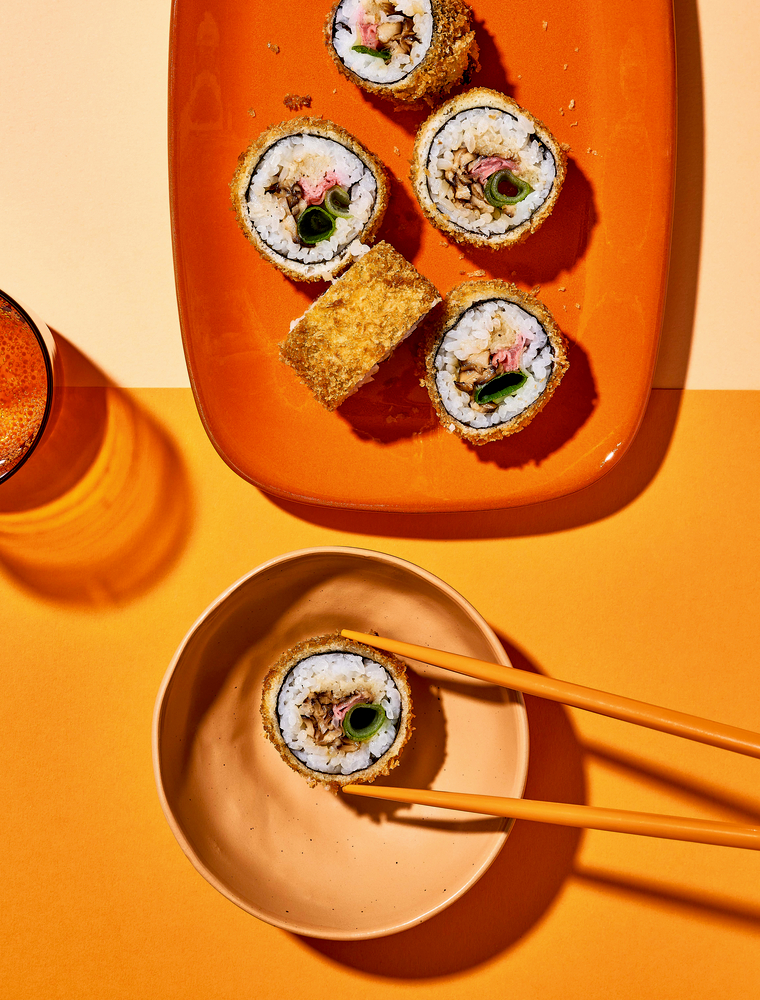 Fünf frittierte Sushistücke auf einem orangefarbenen Teller, eines wird mit Stäbchen über eine beige Schüssel gehoben.