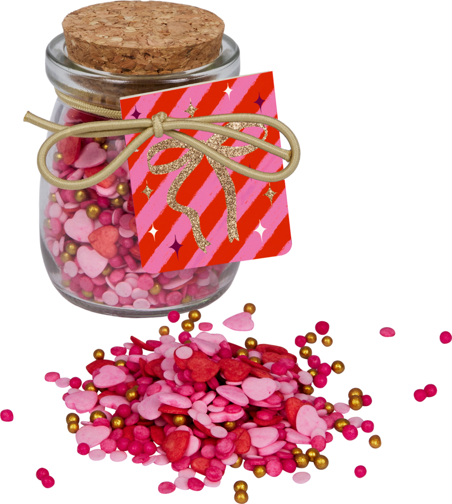 Glas mit Korkdeckel und Anhänger, gefüllt mit rosa, roten und goldenen Zuckerstreuseln in Herz- und Kugelform, teils vor dem Glas verstreut.