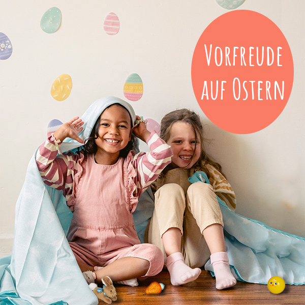 Zwei lachende Mädchen sitzen auf dem Fußboden. Wand mit Ostereiern dekoriert.